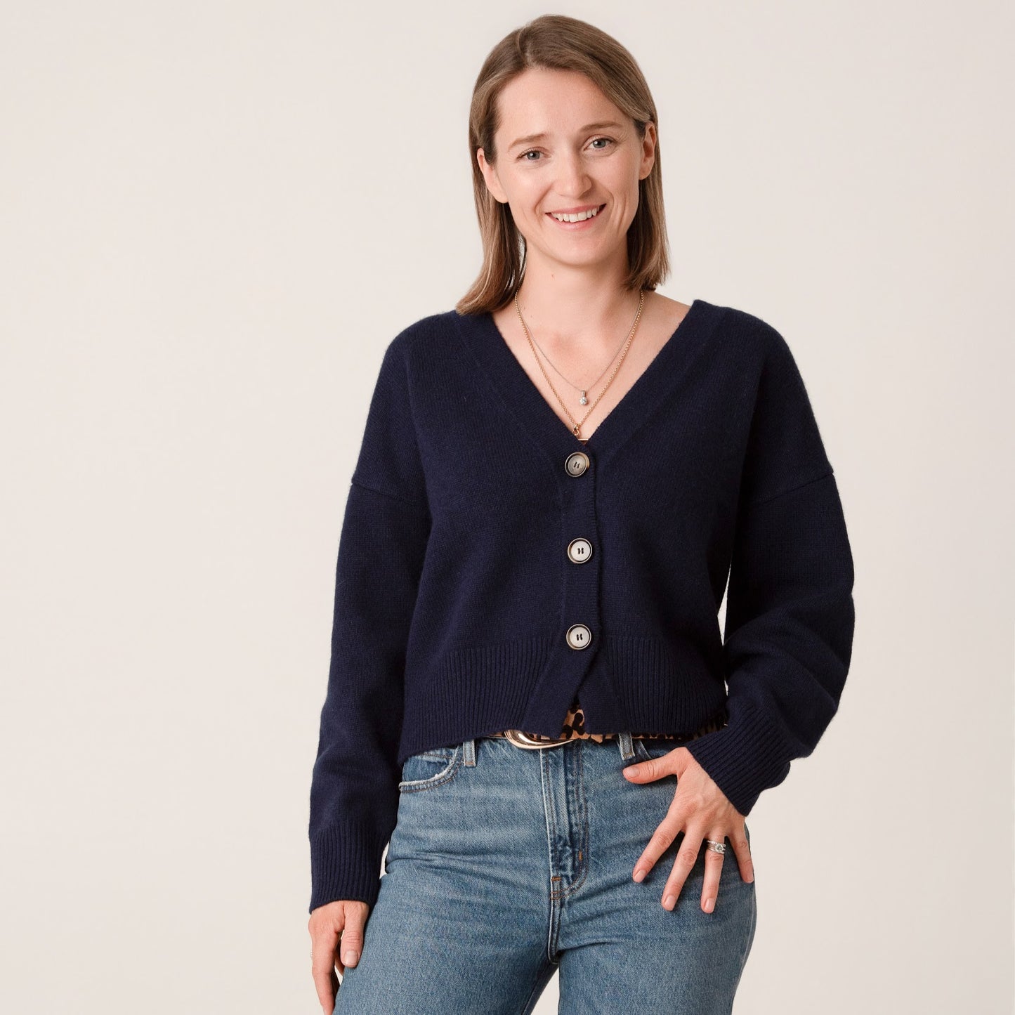 Woman wearing a navy blue cardigan with tortoiseshell buttons and blue jeans on a beige background